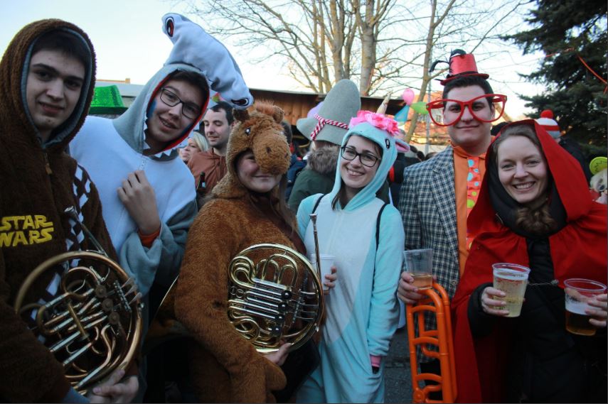 Eine Gruppe von Menschen in verschiedenen Kostümen und Outfits der Stadtkapelle Leonding, einige halten Musikinstrumente und Getränke in den Händen, lächeln gemeinsam bei einer Veranstaltung im Freien.