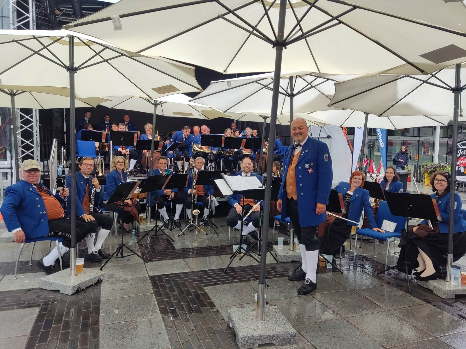 Eine in blauen Uniformen gekleidete Musikkapelle, Stadtkapelle Leonding, tritt im Freien unter großen Sonnenschirmen auf.